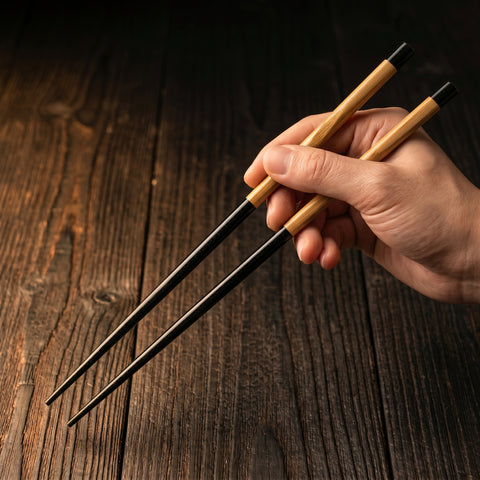 Hand holding a pair of black and wooden chopsticks on a wooden surface